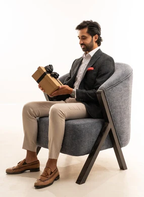 The concept of gratitude and appreciation, expressing love, respect, and admiration, giving gifts on holidays and special occasions, a portrait of a young Arab Gulf Saudi man wearing formal attire sitting on a comfortable gray sofa holding a gift box in his hands, the concept of elegance and beauty, white background.