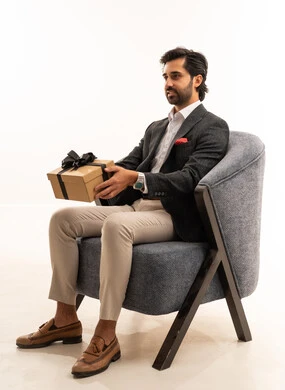 التعبير عن الحب والتقدير والاحترام، مفهوم الشكر والامتنان، تقديم الهدايا بالاعياد والمناسبات الخاصة، بورتريه لشاب عربي خليجي سعودي يرتدي الطقم الرسمي يجلس على اريكة مريحة باللون الرمادي يحمل بيديه صندوق هدية، مفهوم الاناقة والجمال، خلفية بيضاء