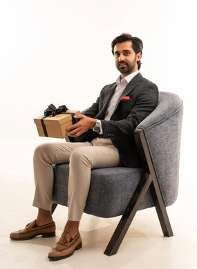 تقديم الهدايا بالاعياد والمناسبات الخاصة، التعبير عن الحب والتقدير والاحترام، مفهوم الشكر والامتنان، بورتريه لشاب عربي خليجي سعودي يرتدي الطقم الرسمي يجلس على كرسي مريح باللون الرمادي يحمل بيديه صندوق هدية، مفهوم الاناقة والجمال، خلفية بيضاء