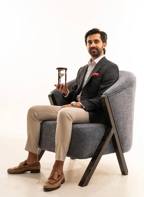 مفهوم إدارة الأعمال وتنظيم الجدول الزمني, إدراك قيمة اللحظات الثمينة, القيام بمراقبة توقيت الرمل, بورتريه لرجل عربي خليجي سعودي يرتدي البدلة الرسمية يجلس على كرسي مريح يمسك بيده الساعة الرملية الزجاجية (البنكام) , خلفية بيضاء