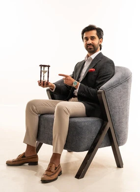 مفهوم إدارة الأعمال وتنظيم الجدول الزمني, إدراك قيمة اللحظات الثمينة, القيام بمراقبة توقيت الرمل, بورتريه لرجل عربي خليجي سعودي يرتدي البدلة الرسمية يجلس على كرسي مريح يمسك بيده الساعة الرملية الزجاجية (البنكام) , خلفية بيضاء
