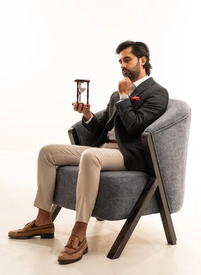 إدراك قيمة اللحظات الثمينة, القيام بمراقبة توقيت الرمل, مفهوم إدارة الأعمال وتنظيم الجدول الزمني, بورتريه لسيد عربي خليجي سعودي يرتدي البدلة الرسمية يجلس على كرسي مريح يمسك بيده الساعة الرملية الزجاجية (البنكام) يضع يده على ذقنه بتعابير التفكير, خلفية بيضاء