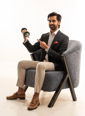 Physical activity to maintain health and physical fitness, exercising with dumbbells, a portrait of an Arab Gulf Saudi businessman wearing a formal suit sitting on a comfortable chair lifting weights (dumbbells), attention to appearance, white background.