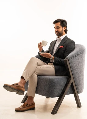 مفهوم الاناقة والرجولة, بورتريه لرجل أعمال عربي خليجي سعودي يرتدي البدلة الرسمية يجلس على كرسي مريح  يحمل كوب قهوة أبيض وطبق صغير تعابير وجهه هادئة ومبتسمة, الاهتمام بالمظهر الخارجي، خلفية بيضاء