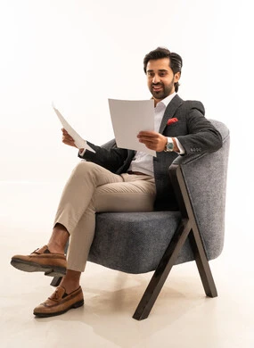 التدقيق في قراءة الملاحظات، اعداد التقارير الخاصة بالعمل، بورتريه لرجل عربي خليجي سعودي يرتدي البدلة الرسمية يجلس على كرسي مريح يحمل بيده مجموعة من الاوراق البيضاء، مراجعة ومتابعة الخطط والمشاريع المكتبية بإيماءات التركيز، خلفية بيضاء