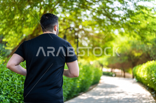 زاوية من الخلف لرجل عربي خليجي سعودي يمارس الرياضة الصباحية في الهواء الطلق ، ممارسة رياضة الجري ، ممارسة الرياضة على طريق الممشى المخصص للمشي بإحدى حدائق السعودية ، نشاط بدني للمحافظة على اللياقة والصحة الجيدة ، ممارسة التمارين الرياضية