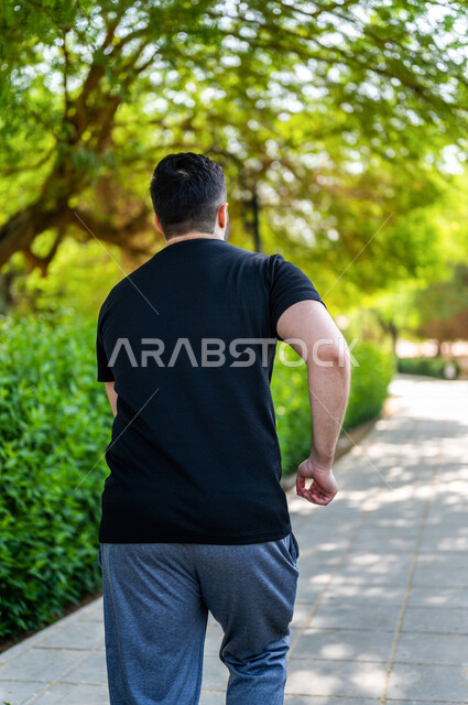 زاوية من الخلف لرجل عربي خليجي سعودي يمارس الرياضة الصباحية في الهواء الطلق ، ممارسة رياضة الجري ، ممارسة الرياضة على طريق الممشى المخصص للمشي بإحدى حدائق السعودية ، نشاط بدني للمحافظة على اللياقة والصحة الجيدة ، ممارسة التمارين الرياضية