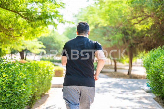 A corner from the back of an Arab man practicing morning sports in the open air, jogging, exercising on the walking path in one of the Saudi gardens, physical activity to maintain fitness and good health, exercising