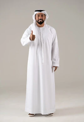Standing with joy, cheerfulness, and happiness, expressing quality and craftsmanship, agreement, satisfaction, and acceptance, a portrait of a young Arab Gulf Emirati man wearing a kandura and ghutrah, raising his thumb with gestures of quality, excellence, and distinction, with a gray background.