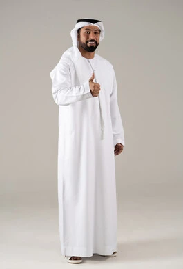 Expressions of joy, cheerfulness, and happiness, the expression of quality and craftsmanship, agreement, satisfaction, and acceptance, a portrait of a young Arab Gulf Emirati man wearing a kandura and ghutrah, raising his thumb with gestures of quality, excellence, and distinction, with a gray background.