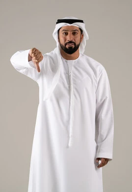 الشعور بالانزعاج من شيء ما,  مفهوم الرفض وعدم القبول، بورترية لرجل عربي خليجي اماراتي يرتدي الكندورة والغترة يشير بإصبع الابهام للاسفل بإيماءات عدم الاعجاب والتقييم السلبي، خلفية رمادية