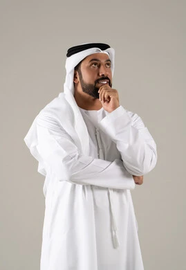 A portrait of an Emirati Gulf Arab man wearing a kandura and ghutrah, resting his hand on his chin with gestures of thinking and distraction, embodying the concepts of listening and focus, aspiration and planning for the future, problem-solving and decision-making, with a gray background.