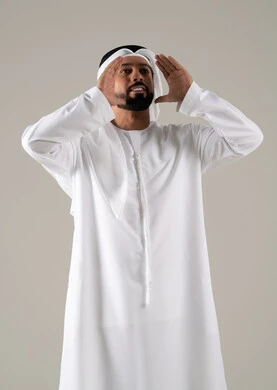 A portrait of an Arab Gulf Emirati man wearing a kandura and ghutrah, placing his hand on his mouth in gestures of calling someone, chatting, conversations, and social communication, calling and speaking loudly, with a gray background.