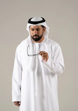 The concept of visual impairment, elegance, and attention to appearance, a portrait of an Emirati Gulf Arab man wearing a kandura, ghutrah, and prescription glasses, with a gray background.