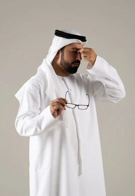The concept of visual impairment, gestures of fatigue and exhaustion, elegance and attention to appearance, a portrait of an Emirati Gulf Arab man wearing a kandura, ghutrah, and prescription glasses, with a gray background.