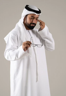 The feeling of pain and headache, the concept of poor eyesight, elegance and attention to appearance, a portrait of an Arab Gulf Emirati man wearing a kandura, ghutrah, and prescription glasses, with a gray background.