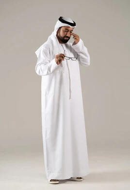 Gestures of fatigue and exhaustion, the concept of poor eyesight, elegance and attention to external appearance, a portrait of an Emirati Gulf Arab man wearing a kandura, ghutrah, and prescription glasses, with a gray background.