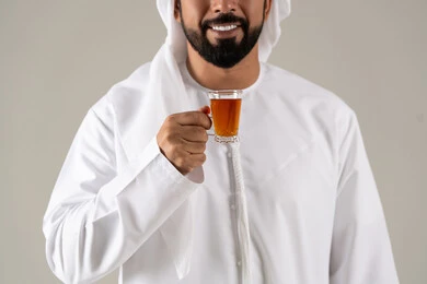 A portrait of an Emirati Gulf Arab man wearing a kandura and ghutrah, holding a cup of tea in his hand, expressions of quality and excellence, enjoying hot beverages, looking with gestures of happiness, the concept of caring about appearance, local Emirati national products, gray background.