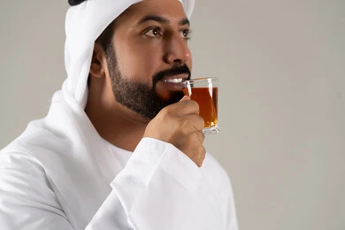 مفهوم الاهتمام بالمظهر الخارجي، منتجات محلية وطنية اماراتيه ، تعابير الجودة والامتياز ، الاستمتاع في تناول المشروبات الساخنه ، النظر بإيماءات السعادة ، بورتريه لرجل عربي خليجي اماراتي يرتدي الكندورة والغترة يحمل بيده كوب من الشاي، خلفية رمادية