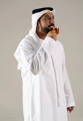 النظر بإيماءات السعادة ، مفهوم الاهتمام بالمظهر الخارجي، تعابير الجودة والامتياز ، الاستمتاع في تناول المشروبات الساخنه ، منتجات محلية وطنية اماراتيه ، بورتريه لرجل عربي خليجي اماراتي يرتدي الكندورة والغترة يحمل بيده كوب من الشاي، خلفية رمادية