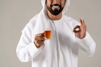 الاستمتاع في تناول المشروبات الساخنه ،تعابير الجودة والامتياز ،  النظر بإيماءات السعادة ، مفهوم الاهتمام بالمظهر الخارجي، منتجات محلية وطنية اماراتيه ، بورتريه لرجل عربي خليجي اماراتي يرتدي الكندورة والغترة يحمل بيده كوب من الشاي، خلفية رمادية