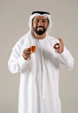 Expressions of quality and excellence, enjoying hot beverages, looking with gestures of happiness, the concept of caring about appearance, locally made Emirati national products, a portrait of an Arab Gulf Emirati man wearing a kandura and ghutrah holding a cup of tea in his hand, gray background.