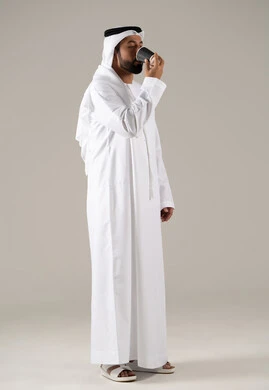 Saudi national local products, the concept of caring about appearance, gestures of enjoyment and integration, consuming hot beverages, a portrait of an Emirati Gulf Arab man wearing a kandura and ghutrah holding a cup of coffee in his hand, gray background.