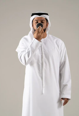 تناول المشروبات الساخنه ،ايماءات الاستمتاع والاندماج، مفهوم الاهتمام بالمظهر الخارجي، منتجات محلية وطنية سعودية، بورتريه لرجل عربي خليجي اماراتي يرتدي الكندورة والغترة يحمل بيدها كوب من القهوة، خلفية رمادية
