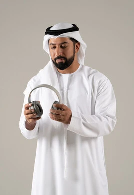 مفهوم التسليه والترفيه, استخدام تقنية الهيدفون، الاستماع الى الأغاني والموسيقى ، بورتريه لرجل عربي خليجي اماراتي يرتدي الكندورة والغترة يضع سماعة الرأس يقوم بالإصغاء والاستماع إلى شيء ما، خلفية رمادية