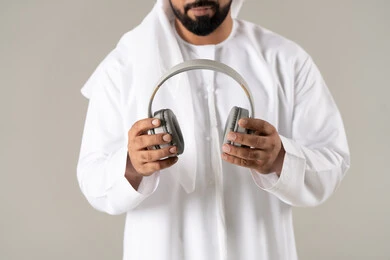 بورتريه لرجل عربي خليجي اماراتي يرتدي الكندورة والغترة يضع سماعة الرأس يقوم بالإصغاء والاستماع إلى شيء ما، استخدام تقنية الهيدفون، الاستماع الى الأغاني والموسيقى ، خلفية رمادية