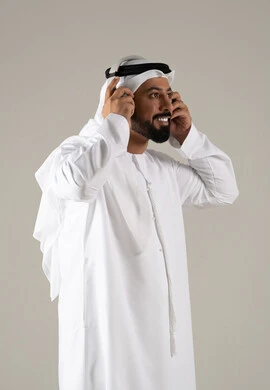 استخدام تقنية الهيدفون، الاستماع الى الأغاني والموسيقى ، بورتريه لرجل عربي خليجي اماراتي يرتدي الكندورة والغترة يضع سماعة الرأس يقوم بالإصغاء والاستماع إلى شيء ما، خلفية رمادية