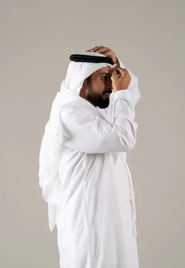 مفهوم الرجولة والثقة بالنفس, بورتريه لرجل عربي خليجي إماراتي يرتدي الكندورة ويعدل من وضعية الغترة البيضاء ينظر بتعابير السرور، الأناقة والاهتمام بالمظهر الخارجي، خلفية رماديه.