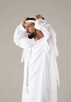 A portrait of an Emirati Gulf Arab man wearing a kandura and adjusting the position of his white ghutrah, looking with expressions of joy, elegance, and attention to appearance, embodying the concept of masculinity and self-confidence, with a gray background.