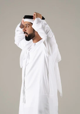 مفهوم الرجولة والثقة بالنفس, بورتريه لرجل عربي خليجي إماراتي يرتدي الكندورة ويعدل من وضعية الغترة البيضاء ينظر بتعابير السرور، الأناقة والاهتمام بالمظهر الخارجي، خلفية رماديه.