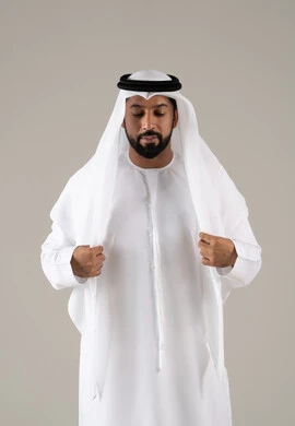 Elegance and attention to appearance, the concept of masculinity and self-confidence, a portrait of an Arab Gulf Emirati man wearing a kandura and adjusting the position of his white ghutrah, looking with expressions of joy, with a gray background.