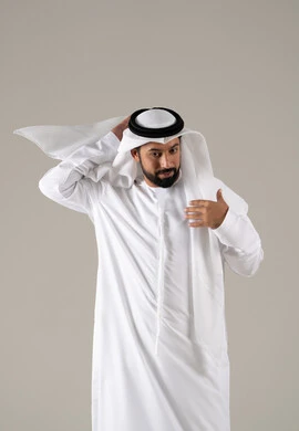 The concept of masculinity and self-confidence, a portrait of an Emirati Gulf Arab man wearing a kandura and adjusting the position of his white ghutrah, looking with expressions of joy, elegance, and attention to appearance, with a gray background.