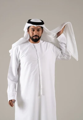 The concept of masculinity and self-confidence, a portrait of an Emirati Gulf Arab man wearing a kandura and adjusting the position of his white ghutrah, looking with expressions of joy, elegance, and attention to external appearance, with a gray background.