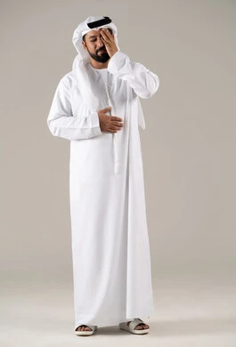 A portrait of an Emirati Gulf Arab man wearing a kandura and ghutrah, placing his hands on his head and closing his eyes with gestures of severe headache pain, the feeling of a migraine and tension headache, the sensation of fatigue and loss of concentration, stress and psychological pressure, with a gray background.