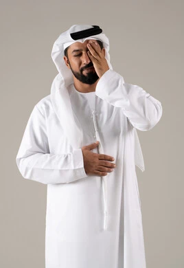 Stress and psychological pressure, the feeling of migraines and headaches, the sensation of fatigue and loss of concentration, a portrait of an Emirati Gulf Arab man wearing a kandura and ghutrah, placing his hands on his head and closing his eyes with gestures of severe headache pain, gray background.