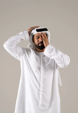 Feeling exhausted and losing focus, experiencing tightness and migraines, stress and psychological pressure, a portrait of an Emirati Gulf Arab man wearing a kandura and ghutrah, placing his hands on his head and closing his eyes with gestures of severe headache, gray background.