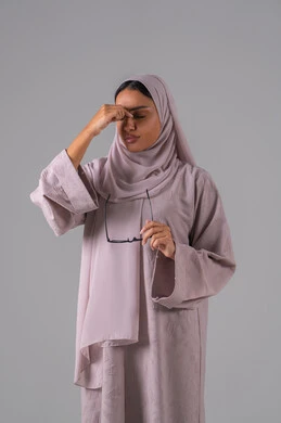 الشعور بالصداع والالم، مفهوم ضعف البصر، الاناقة والاهتمام بالمظهر الخارجي، بورتريه لامرأة عربية خليجية سعودية ترتدي العباءه والحجاب والنظارات الطبيه بايماءات التفاعل والاندماج، خلفية رماديه