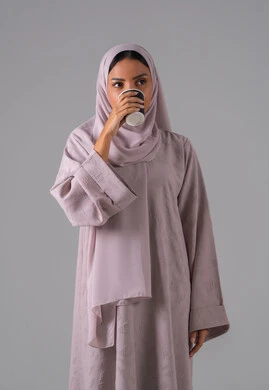 Enjoying hot beverages, looking with gestures of integration, the concept of caring about external appearance, locally made Saudi national products, a portrait of an Arab Gulf Saudi woman wearing an abaya and hijab holding a cup of coffee in her hand, gray background.