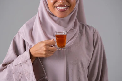 تعابير الجودة والامتياز ، الاستمتاع في تناول المشروبات الساخنه ، النظر بإيماءات السعادة ، مفهوم الاهتمام بالمظهر الخارجي، منتجات محلية وطنية سعودية، بورتريه لامراة عربيه خليجية سعودية ترتدي العباءة والحجاب تحمل بيدها كوب من الشاي، خلفية رمادية