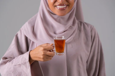 A portrait of a Gulf Arab Saudi woman wearing an abaya and hijab, holding a cup of tea in her hand, with expressions of quality and excellence, enjoying hot beverages, looking with gestures of happiness, the concept of caring about external appearance, local Saudi national products, gray background.