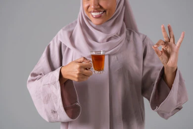 مفهوم الاهتمام بالمظهر الخارجي، منتجات محلية وطنية سعودية، تعابير الجودة والامتياز ، الاستمتاع في تناول المشروبات الساخنه ، النظر بإيماءات السعادة ،بورتريه لامراة عربيه خليجية سعودية ترتدي العباءة والحجاب تحمل بيدها كوب من الشاي، خلفية رمادية