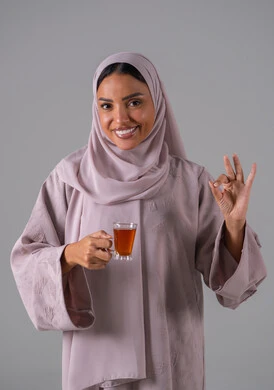 النظر بإيماءات السعادة ، تعابير الجودة والامتياز ، الاستمتاع في تناول المشروبات الساخنه ، مفهوم الاهتمام بالمظهر الخارجي، منتجات محلية وطنية سعودية، بورتريه لامراة عربيه خليجية سعودية ترتدي العباءة والحجاب تحمل بيدها كوب من الشاي، خلفية رمادية
