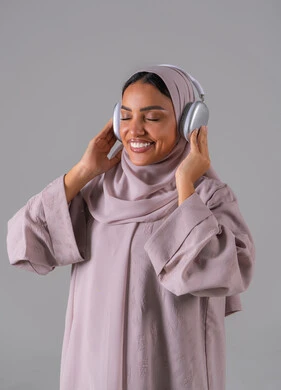 ايماءات السرور والسعادة ،استخدام تقنية الهيدفون، الاستماع الى الأغاني والموسيقى ، بورتريه لامراه عربيه خليجيه سعودية ترتدي العباءة والحجاب تضع سماعة الرأس تقوم بالإصغاء والاستماع إلى شيء ما، خلفية رمادية
