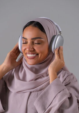 A portrait of a Saudi Arabian Gulf woman wearing an abaya and hijab, wearing headphones, listening to something, using headphone technology, listening to songs and music, with a gray background.