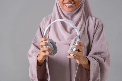 الاستماع الى الأغاني والموسيقى ، استخدام تقنية الهيدفون، بورتريه لامراه عربيه خليجيه سعودية ترتدي العباءة والحجاب تضع سماعة الرأس تقوم بالإصغاء والاستماع إلى شيء ما، خلفية رمادية