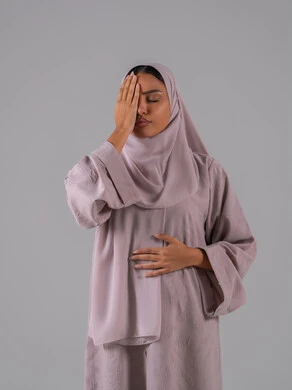 الاجهاد والضغط النفسي, الاحساس بالشقيقية والصداع النصفي, الشعور بالإرهاق وفقدان التركيز, بورتريه لامرأه عربيه خليجيه سعوديه ترتدي العباءة والحجاب تضع يديها على رأسها وتغمض عينيها بإيماءات ألم الرأس الشديد, خلفية رمادية